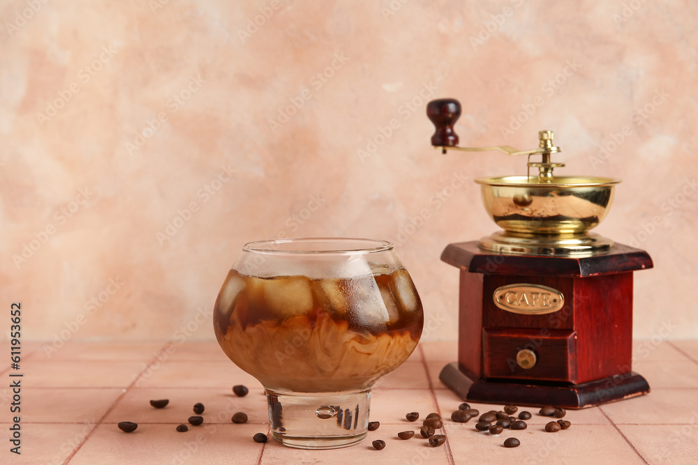 Glass of ice coffee with beans and grinder on beige tile table near wall