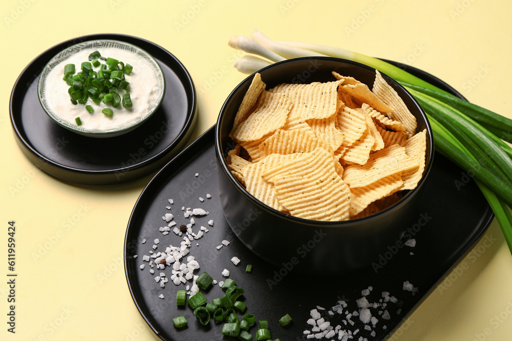 Bowl of tasty sour cream with sliced green onion and potato chips on yellow background