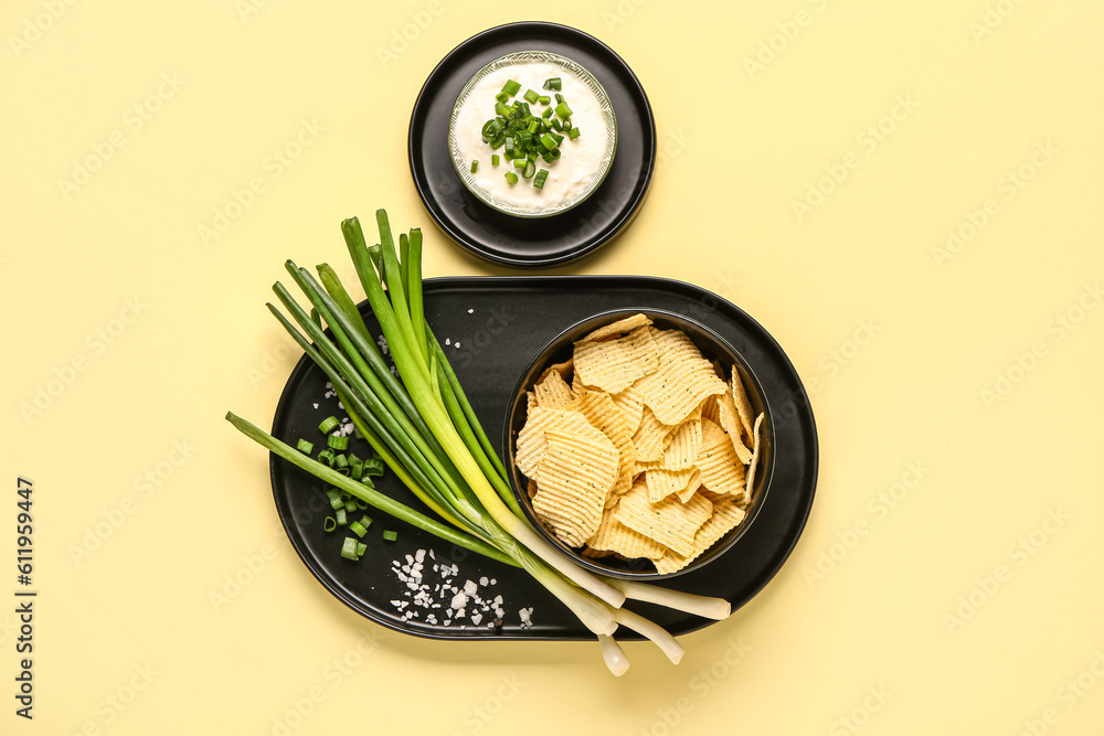 Bowl of tasty sour cream with sliced green onion and potato chips on yellow background