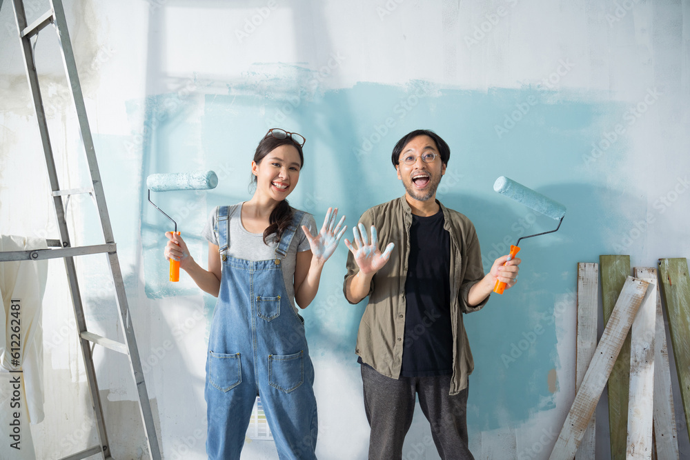 Young Asian couple repairing and painting the wall with blue paint using a roller during renovation 