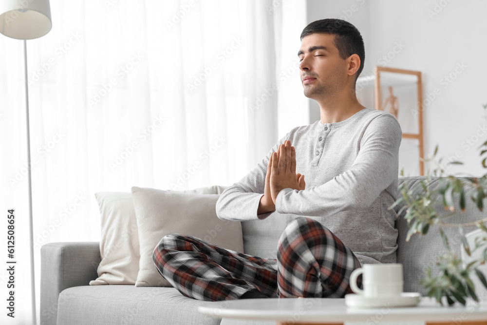 Young man in pajamas meditating on sofa at home