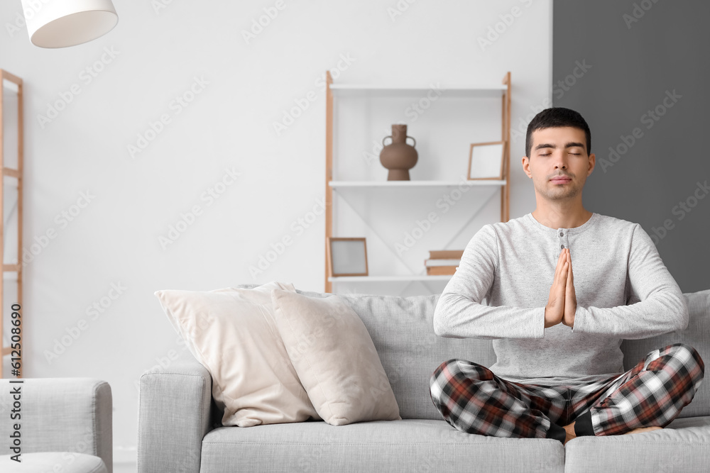 Young man in pajamas meditating on sofa at home