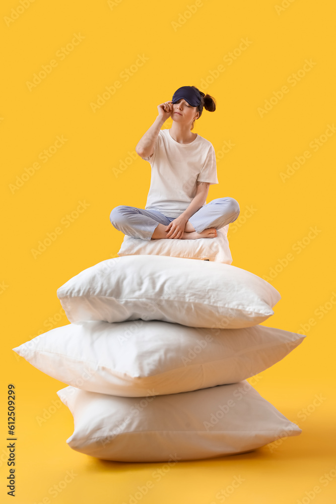 Young woman with sleeping mask and soft pillows sitting on yellow background