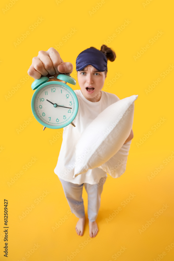 Shocked young woman with sleeping mask, alarm clock and pillow on yellow background