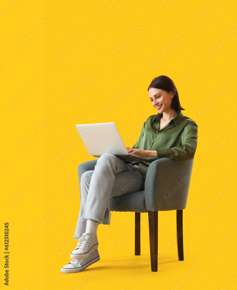 Beautiful woman using laptop in soft armchair on yellow background