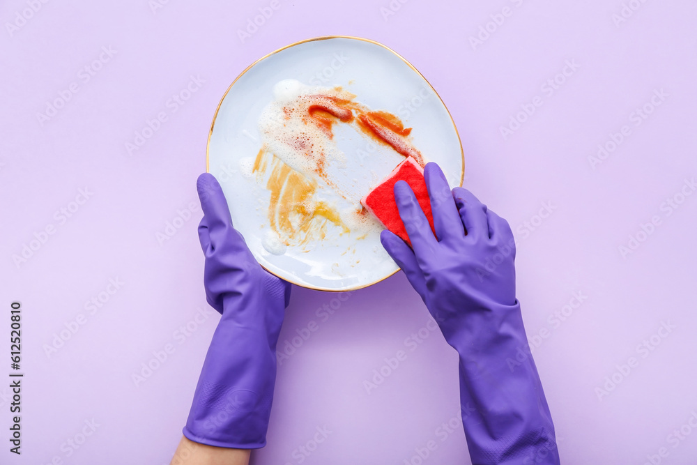 Female hands in rubber gloves washing dirty plate with sponge on lilac background