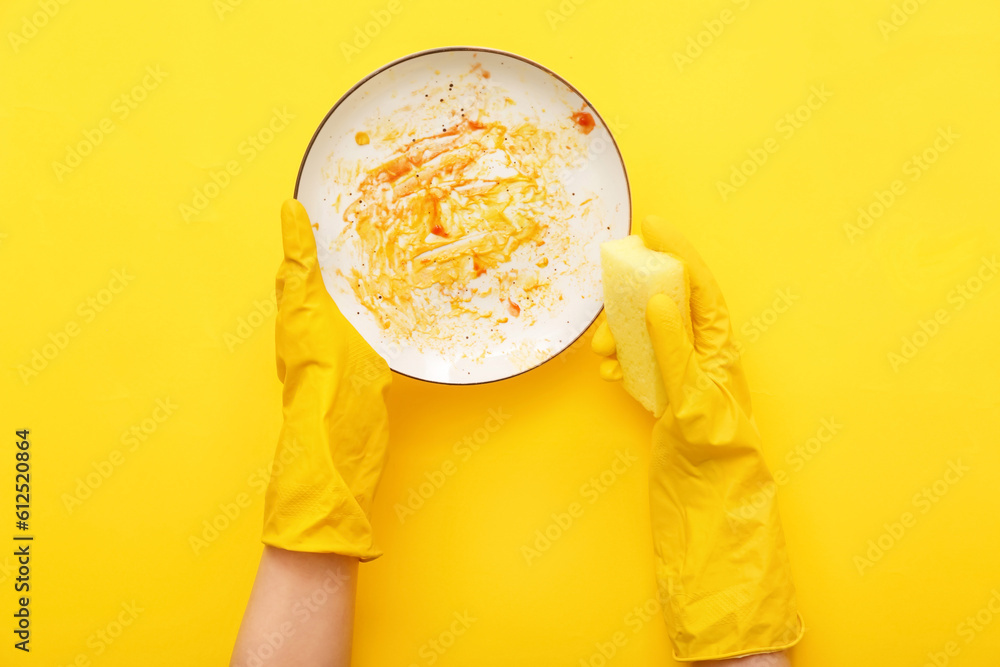 Female hands in rubber gloves washing dirty plate with sponge on yellow background