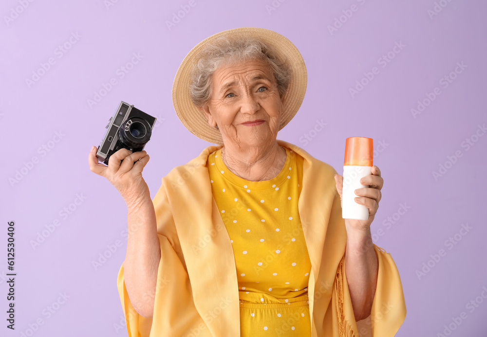 Senior woman with sunscreen cream and photo camera on lilac background