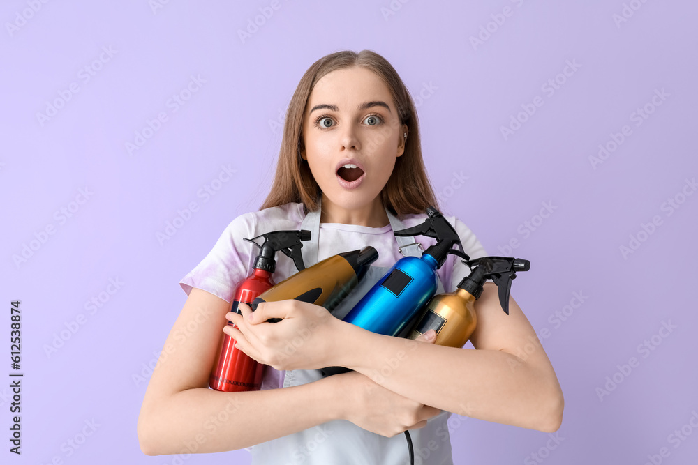 Surprised female hairdresser with sprays and dryer on lilac background