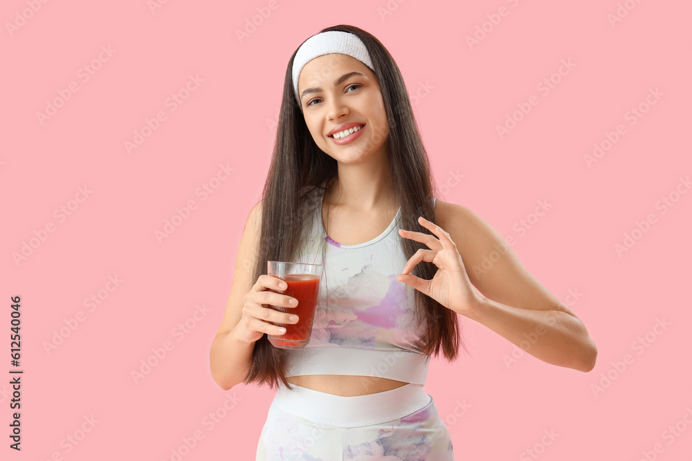 Sporty young woman with glass of vegetable juice showing OK on pink background