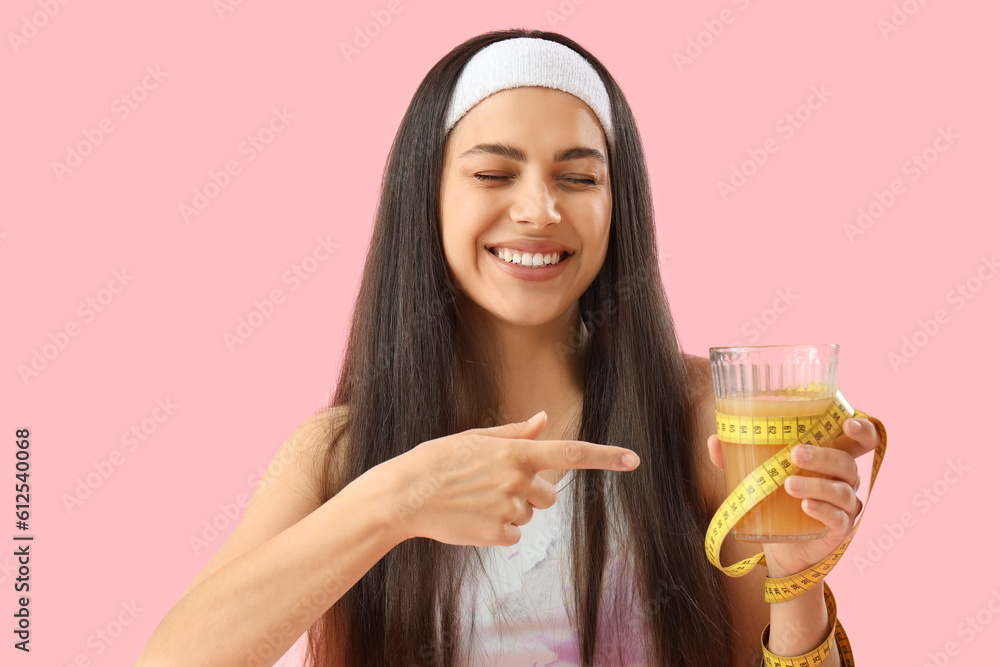 Sporty young woman pointing at glass of vegetable juice and tape measure on pink background