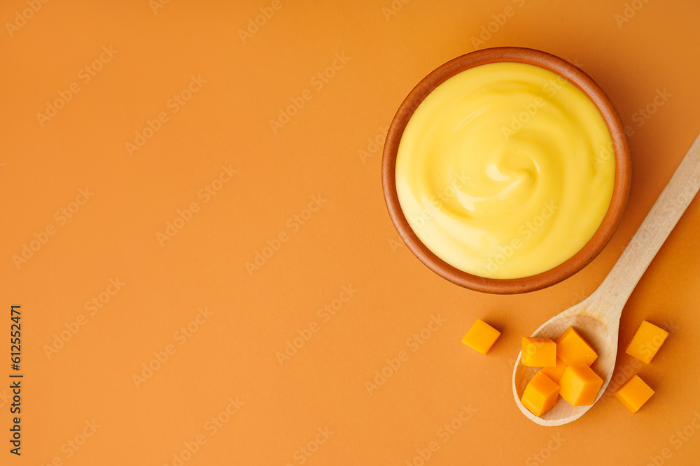 Bowl of tasty cheddar sauce and spoon with cheese cubes on orange background