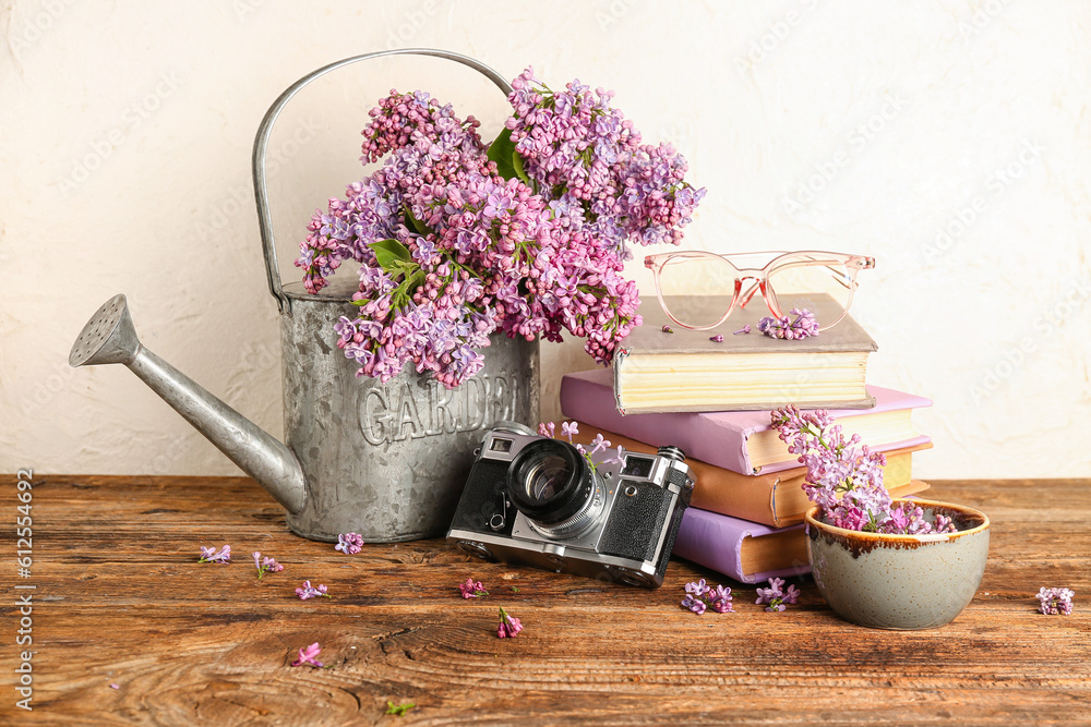 Composition with watering can, camera and lilac twigs on wooden table near white wall