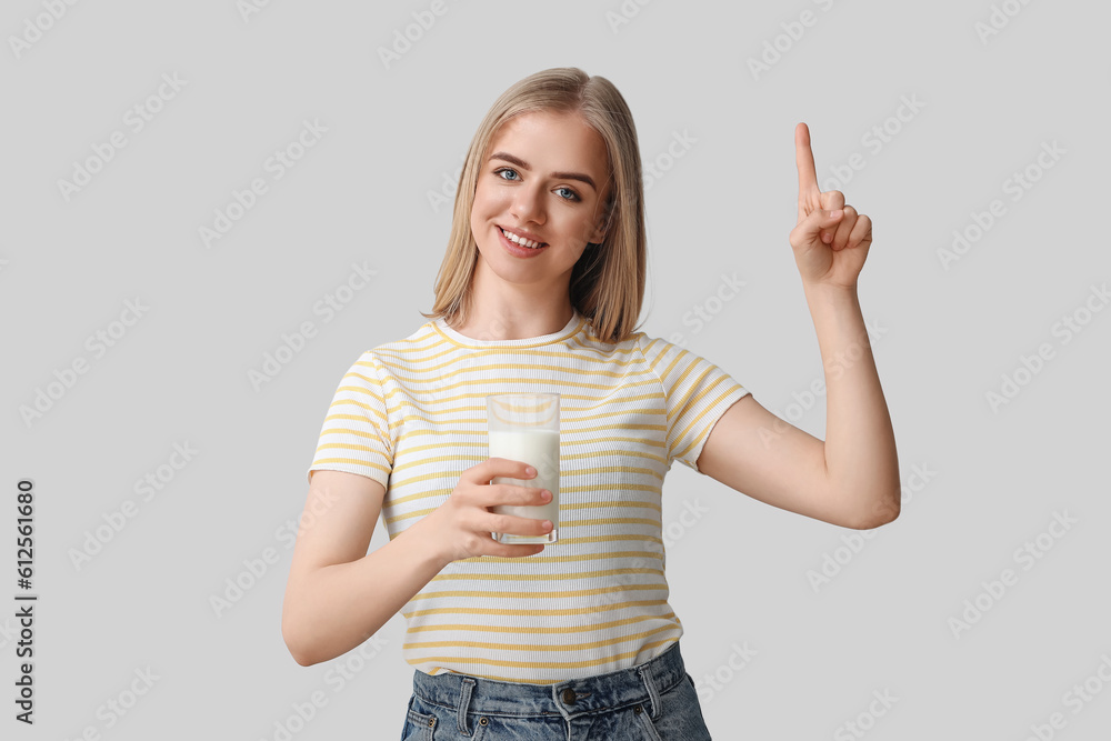 Beautiful young woman with glass of milk pointing at something on grey background