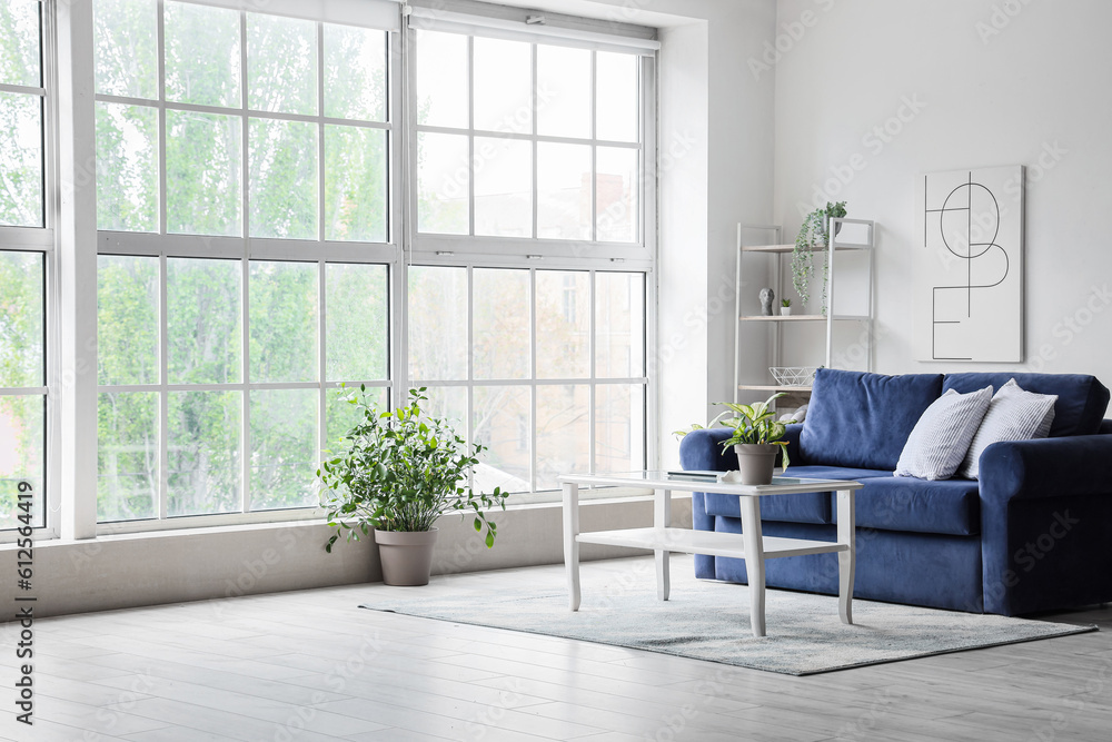 Interior of light living room with cozy blue sofa and modern laptop on table near big window