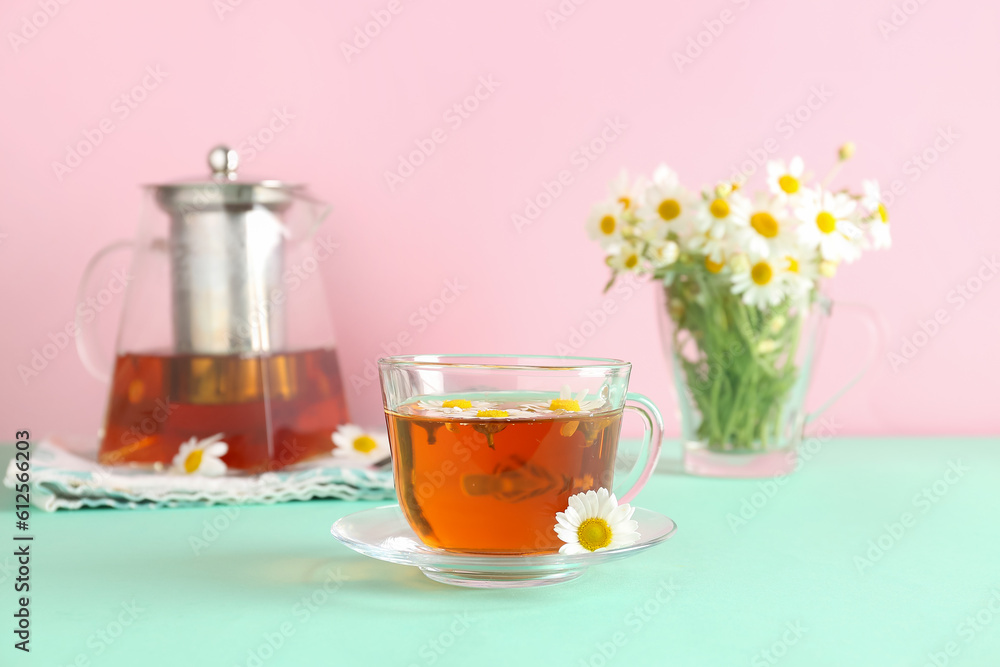 Teapot with cup of natural chamomile tea and flowers on turquoise table near pink wall