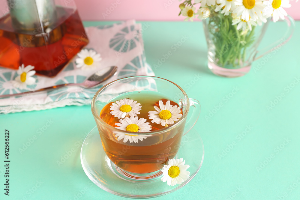 Teapot with cup of natural chamomile tea and flowers on turquoise table near pink wall