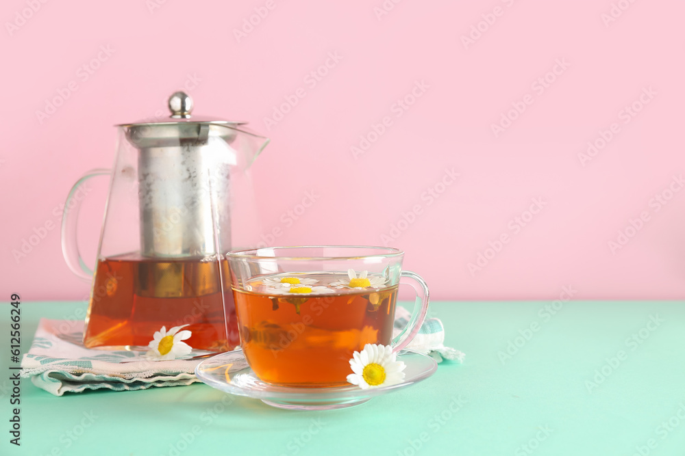 Teapot with cup of natural chamomile tea and flowers on turquoise table near pink wall