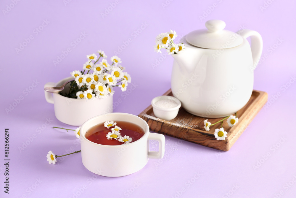 Wooden board with teapot, cups of natural chamomile tea and flowers on lilac background