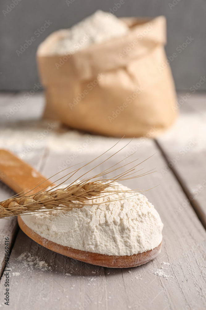 Paper bag with flour, wooden spoon and wheat ear on grey wooden table near wall