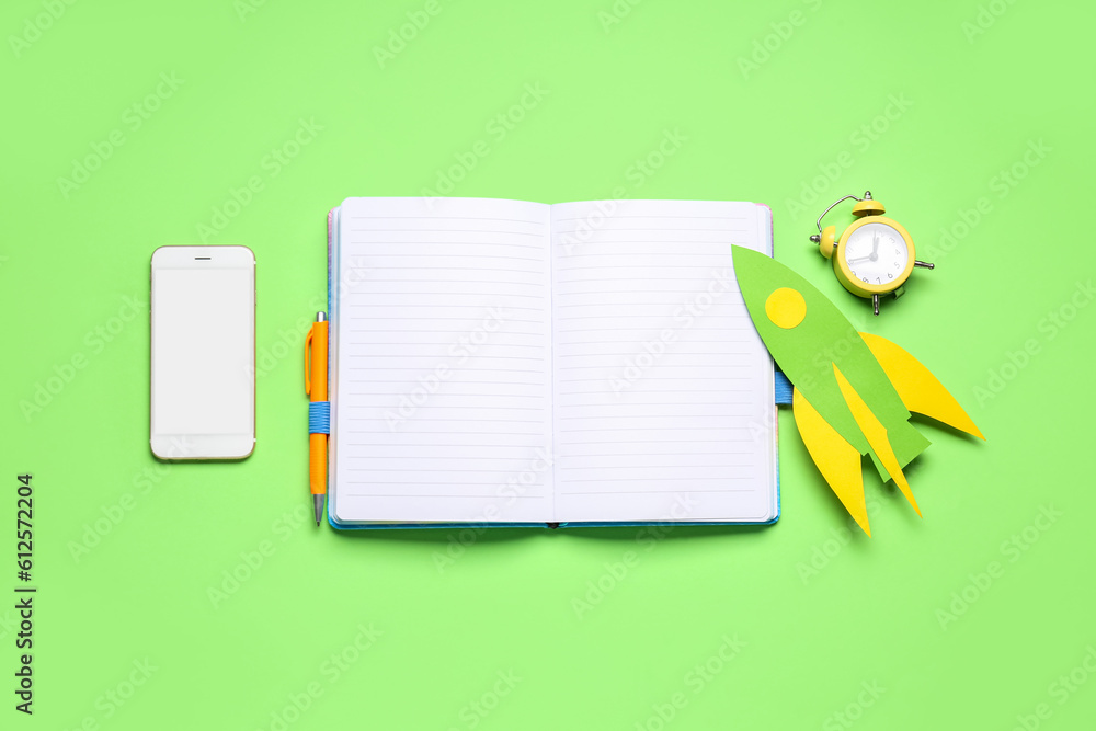 Paper rocket with alarm clock, blank notebook and mobile phone on green background