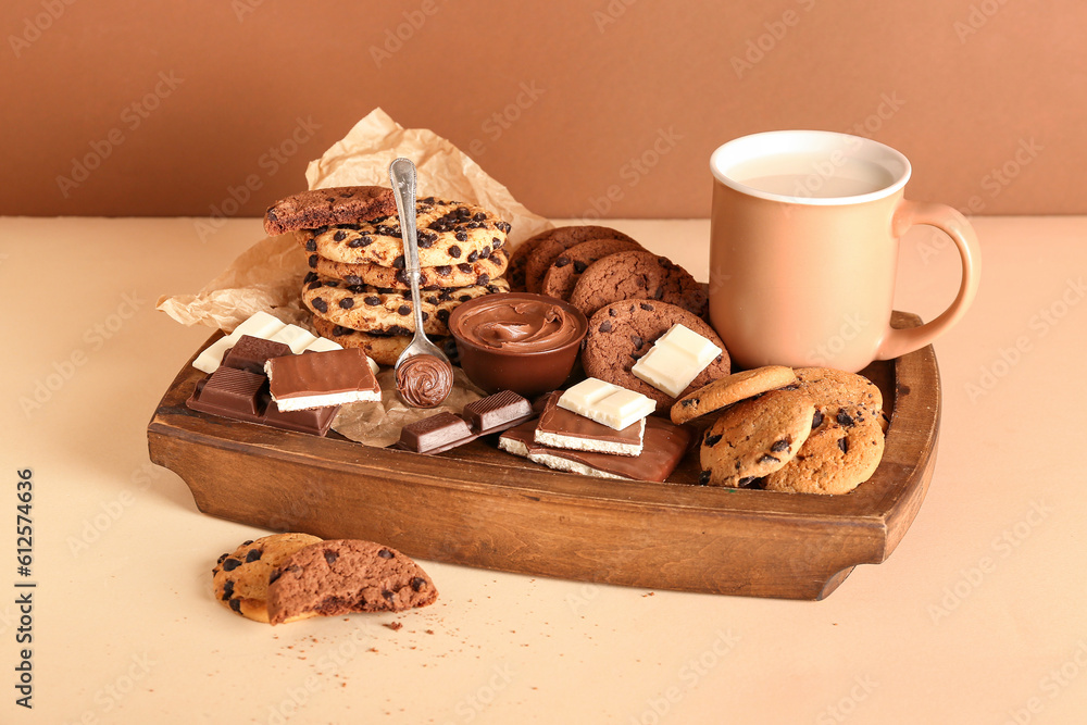 Wooden tray with cookies, chocolate and cup of milk on beige table near brown wall