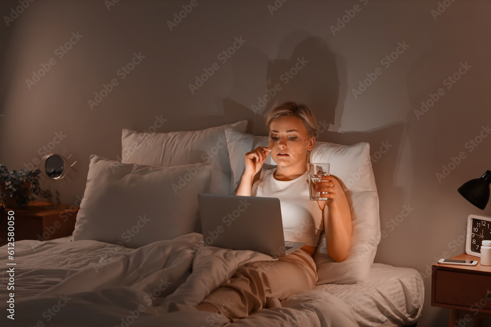 Young woman with laptop taking pill in bed at night