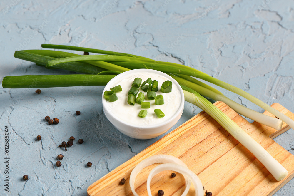 Bowl of tasty sour cream and board with green onion on blue background