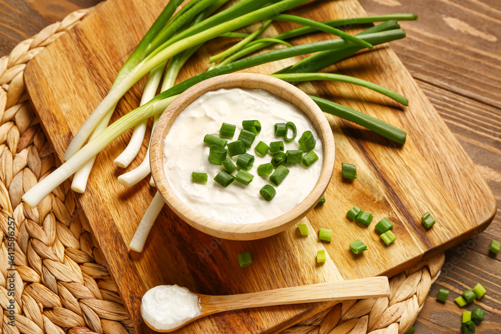 Board and bowl of tasty sour cream with sliced green onion on wooden background