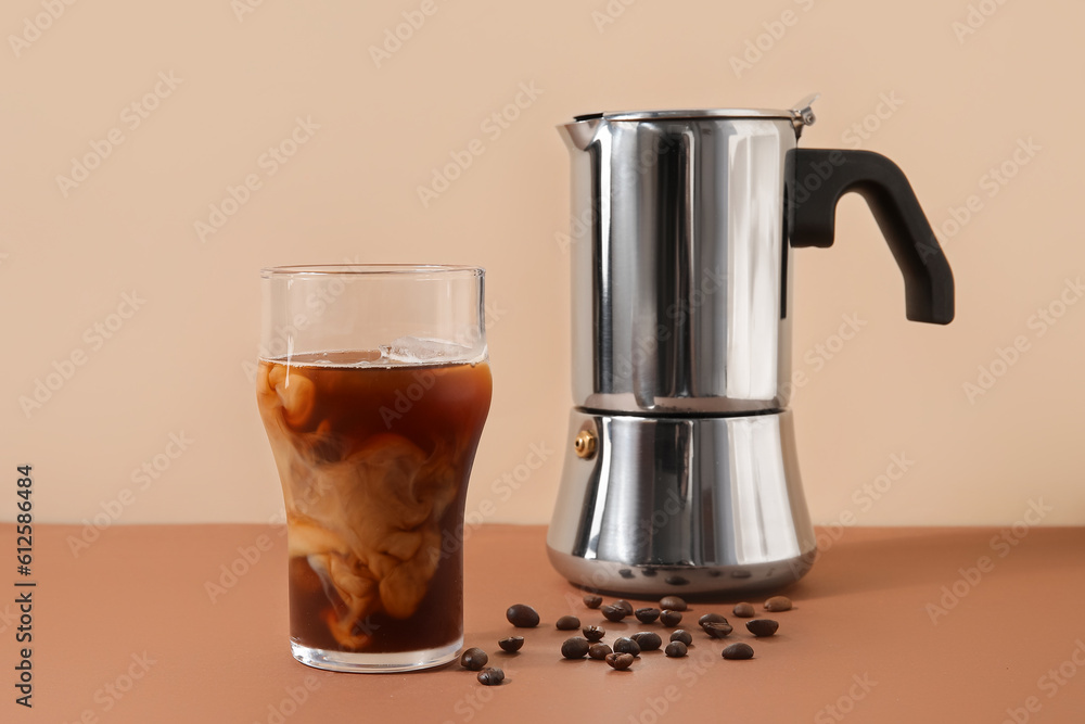 Glass of ice coffee with beans and geyser coffee maker on table near beige wall