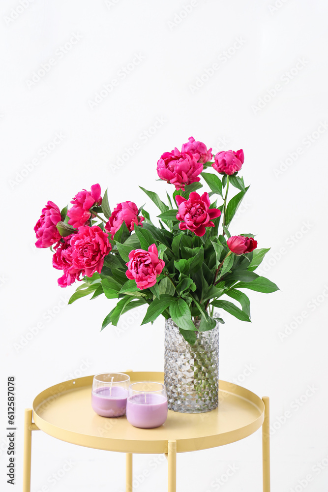 Vase with red peonies and candles on coffee table near white wall