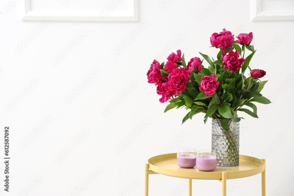 Vase with red peonies and candles on coffee table near white wall