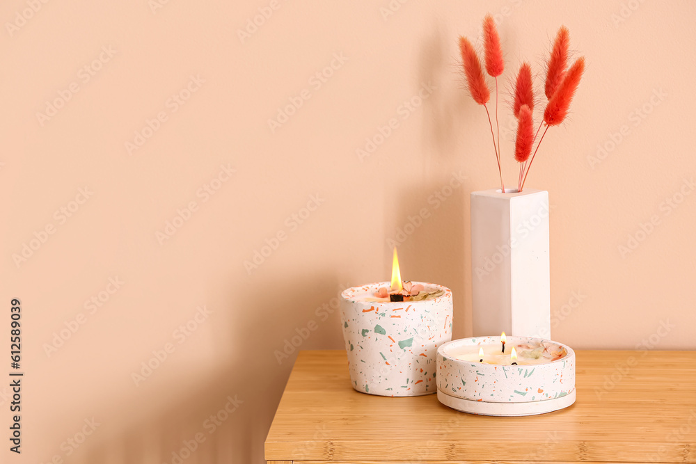 Holders with burning candles and lagurus on table near beige wall in room