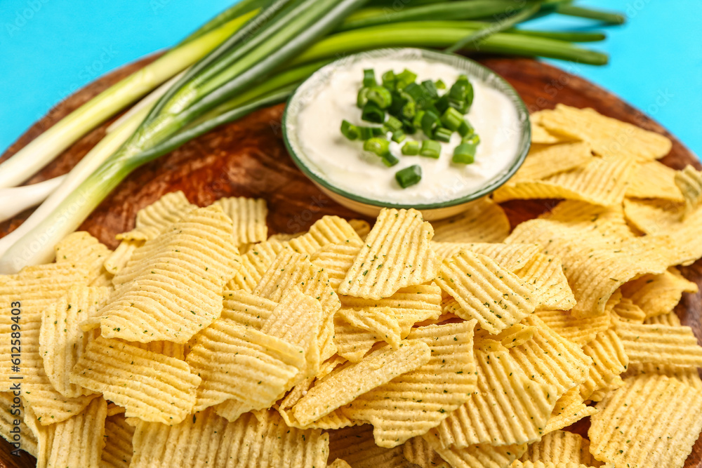 Bowl of tasty sour cream with sliced green onion and potato chips on blue background