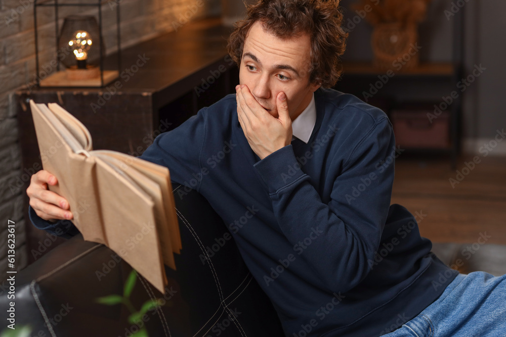 Shocked young man reading book on sofa at home late in evening