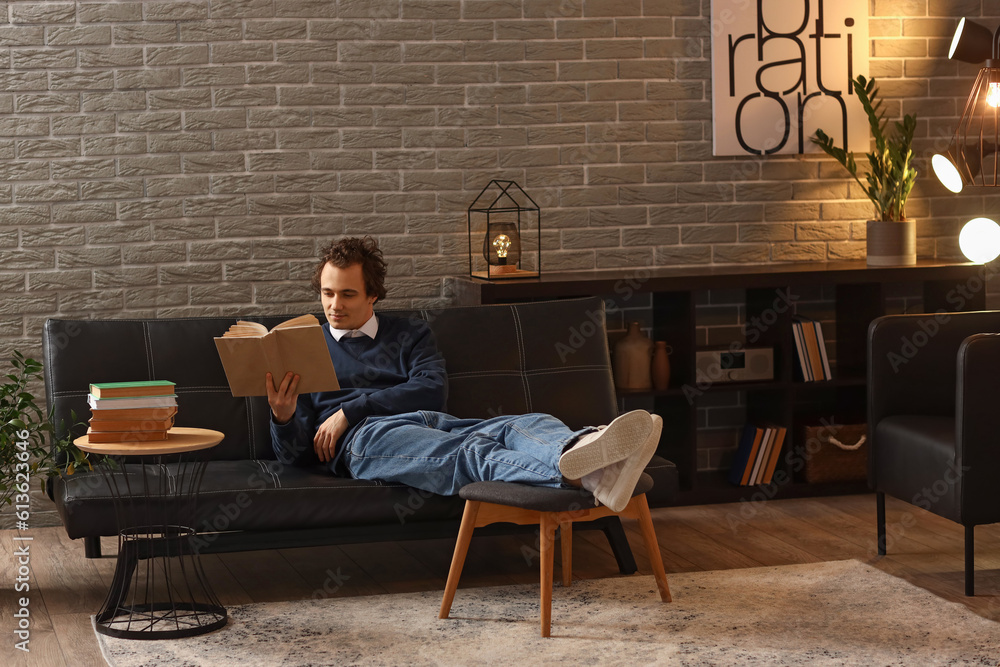 Young man reading book on sofa at home late in evening