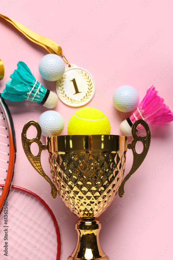 Gold cup with first place medal and sports equipment on pink background, closeup