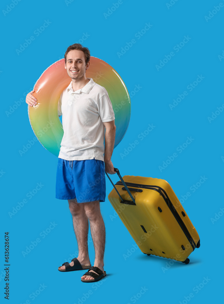 Young man with inflatable ring and suitcase on blue background