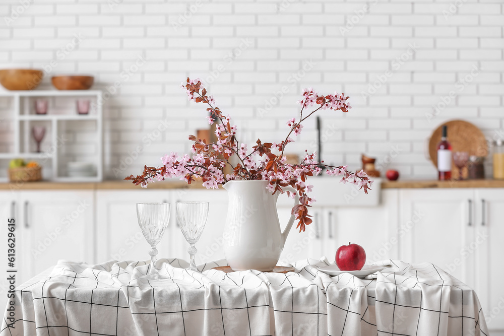 Vase with blooming tree branches, apple and glasses on table in kitchen