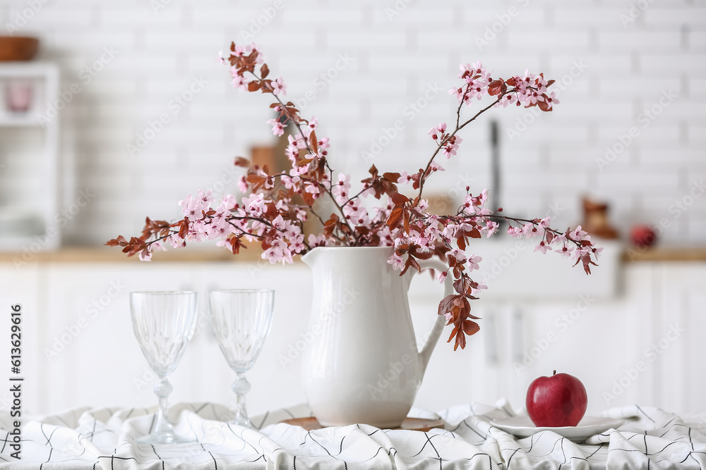 Vase with blooming tree branches, apple and glasses on table in kitchen