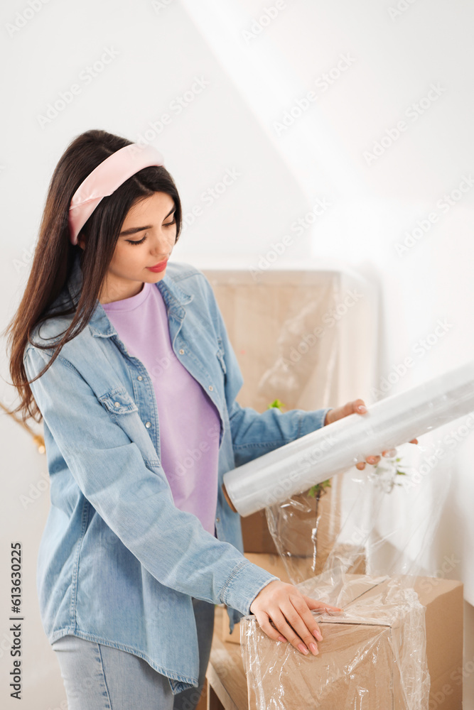 Young woman wrapping cardboard box with stretch film at home on moving day