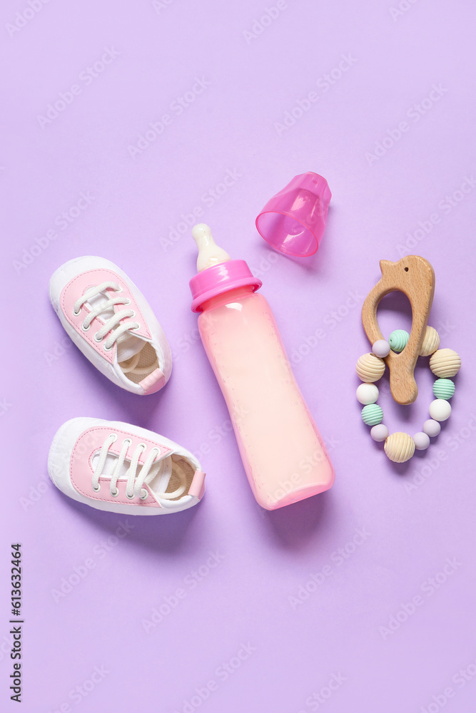 Bottle of milk for baby with rattle and booties on lilac background