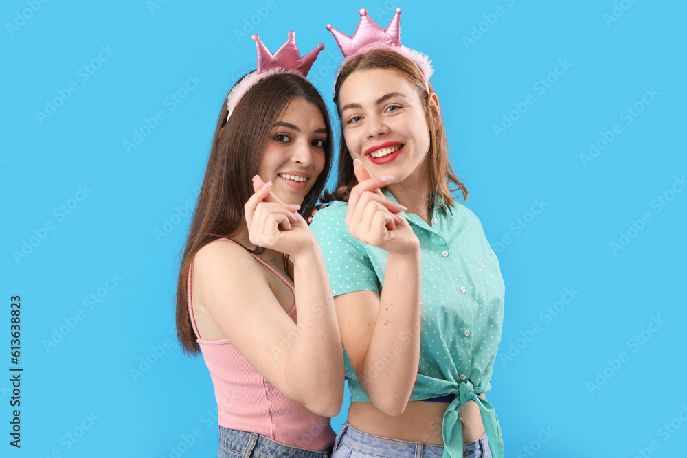 Female friends in crowns making heart shape with their fingers on blue background