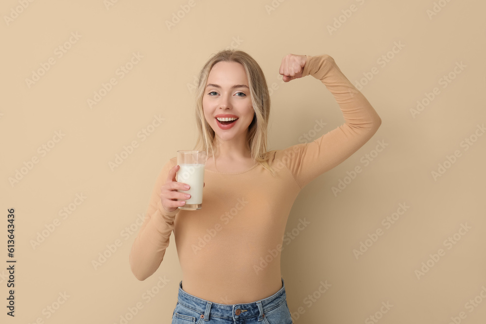 Young woman with glass of milk showing muscles on beige background