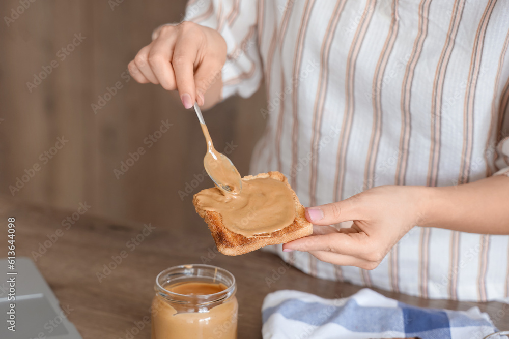 Young woman spreading tasty nut butter onto toast in kitchen, closeup