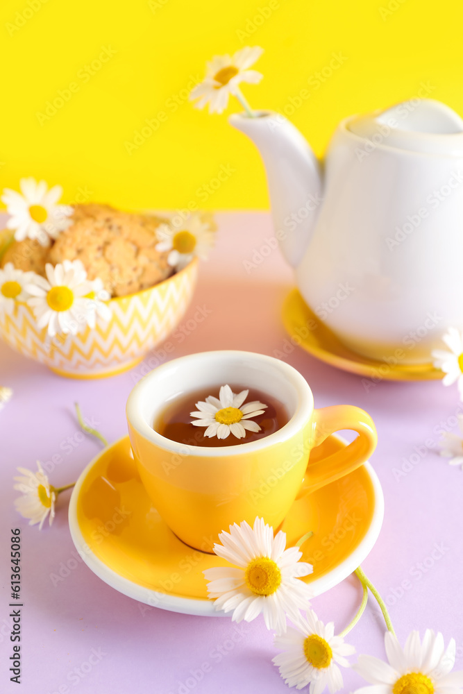 Teapot with cup of natural chamomile tea, cookies and flowers on pink table near yellow wall