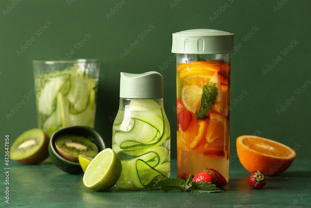 Sports bottles and glass of infused water with different sliced fruits on green background