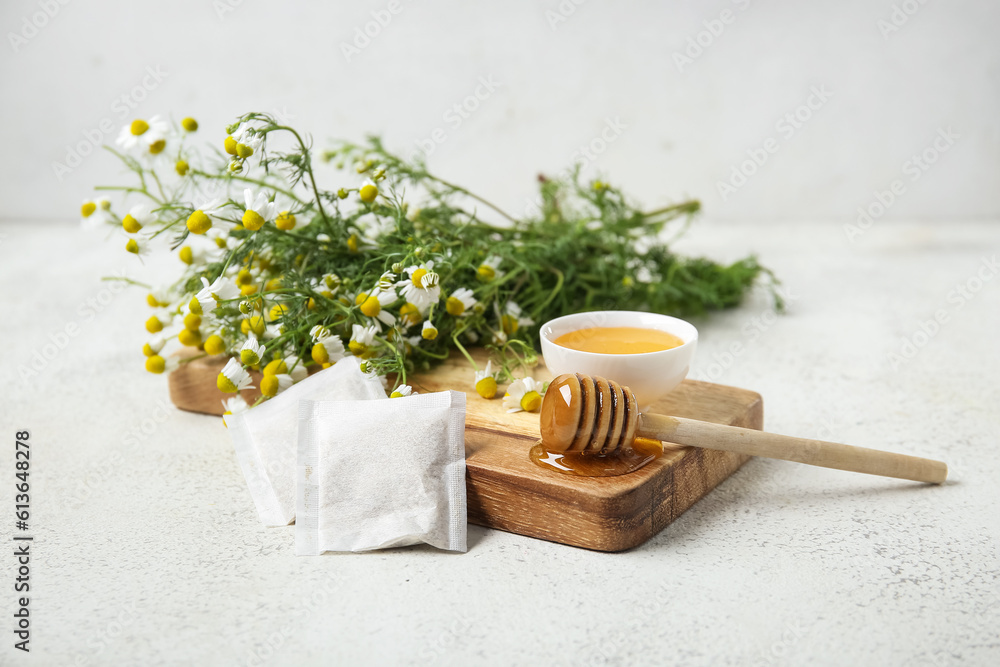 Wooden board with bowl of honey, tea bags and fresh chamomile flowers on light background