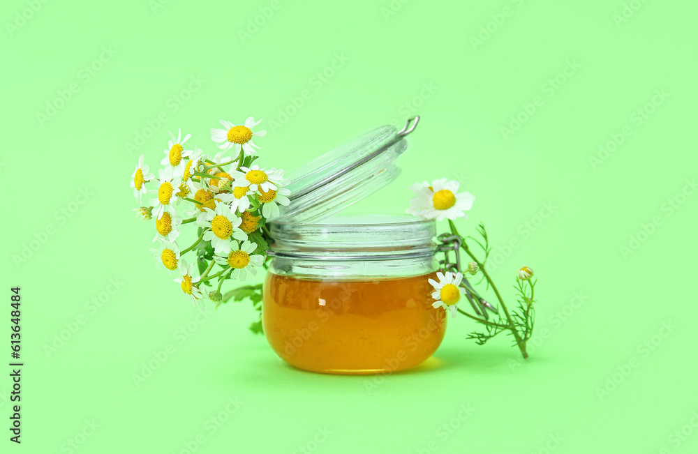 Glass jar of honey and fresh chamomile flowers on green background