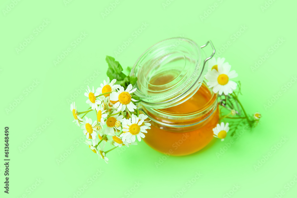 Glass jar of honey and fresh chamomile flowers on green background